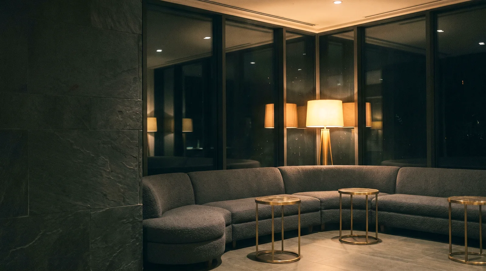 A curved charcoal boucle sectional with brass tables in a hotel lounge at night