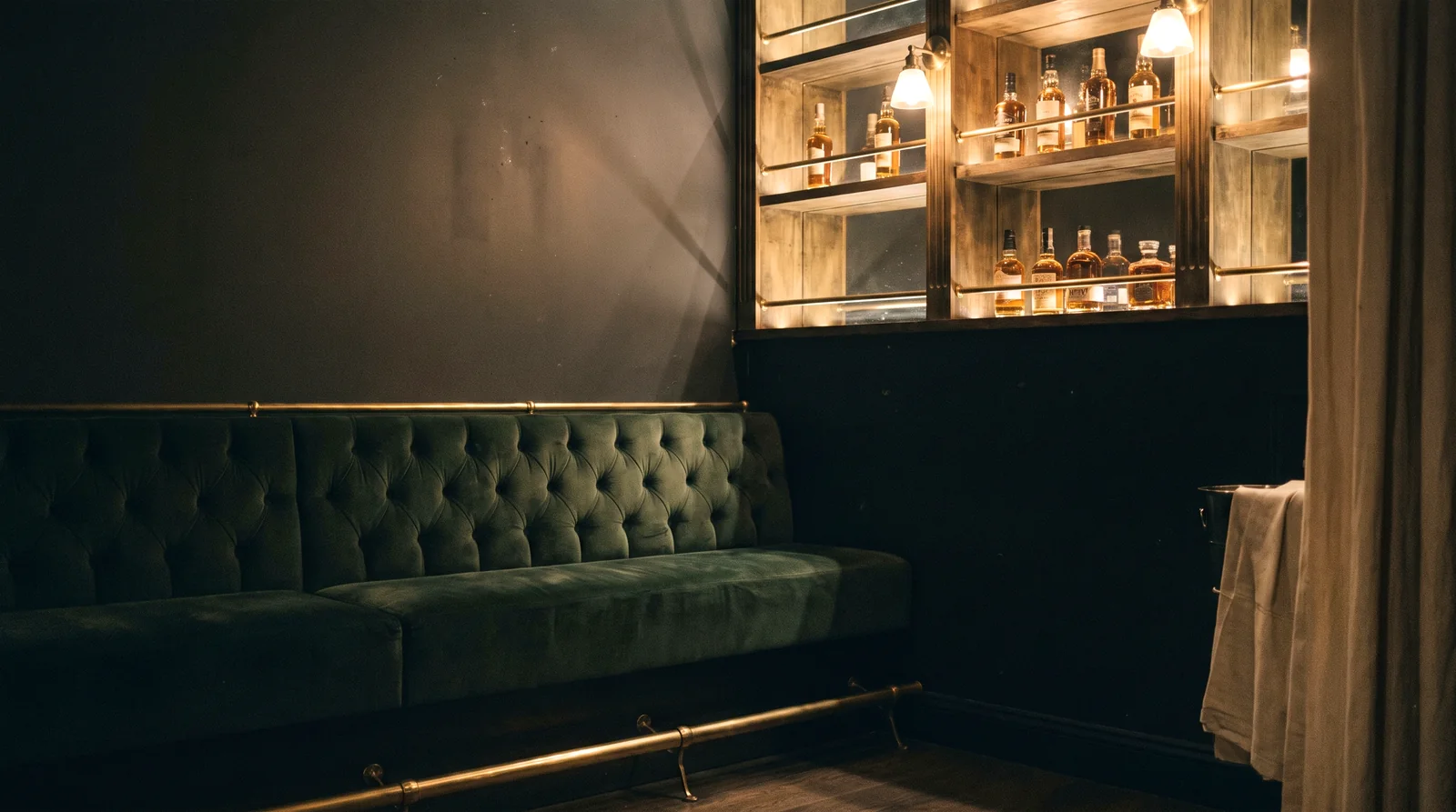A green velvet button-tufted banquette below lit liquor shelving